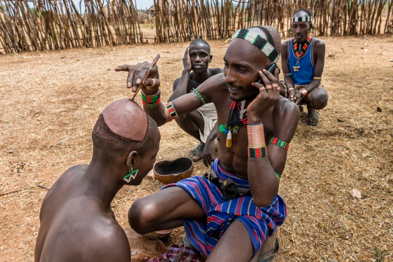 A man from the Hamer tribe decorates his friends head with a tradional clay skull cap while talking on a cell phone.